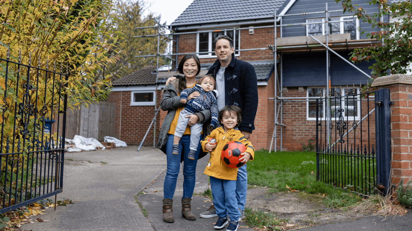 a couple and 2 children (one held by the woman) stand in front of their house which has scaffolding up on outside