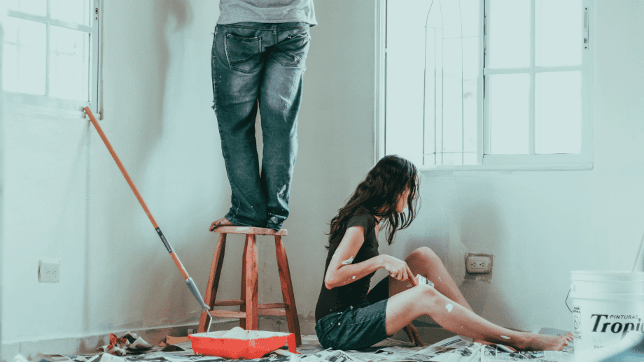 A couple paint a room, one sitting down, and other on a stool