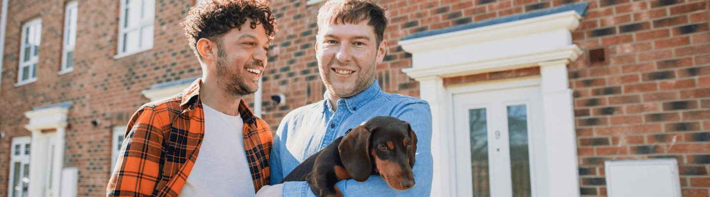 2 men standing outside a house holding a dog